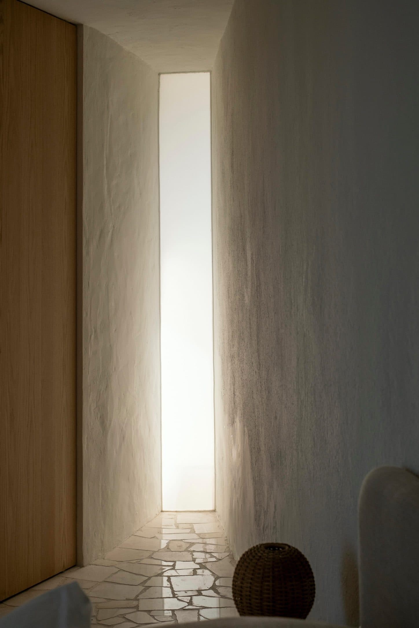 A narrow slit of light in a plaster wall beside a timber panel and a clay vessel.