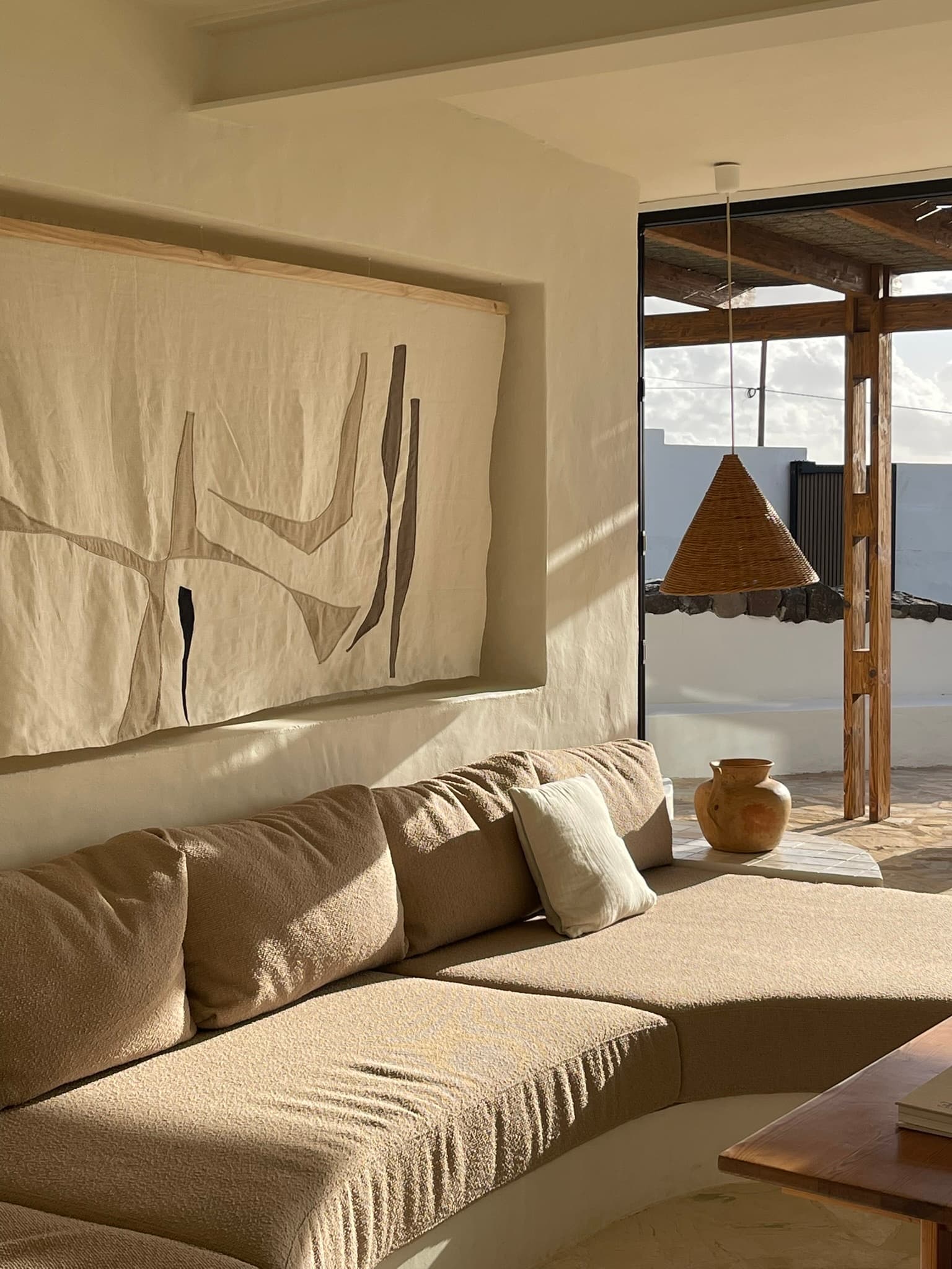 A curved plaster sofa beneath a large woven wall hanging, cushions in stone and clay.