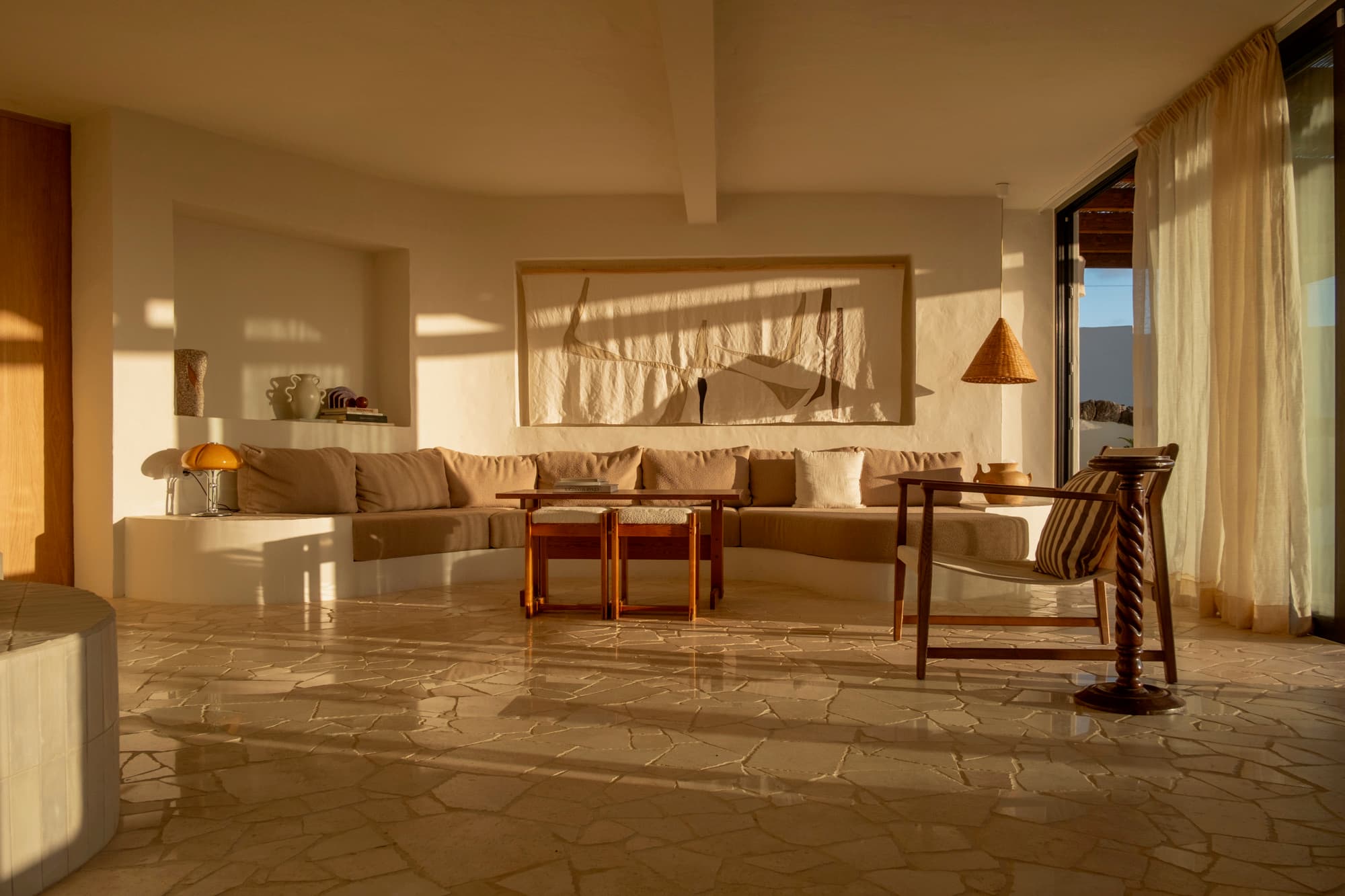 The living room in late golden light: a curved plaster sofa, timber tables and linen curtains.